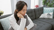 © Krakenimages.com - A contemplative young asian woman sits thoughtfully in a cozy modern living room.