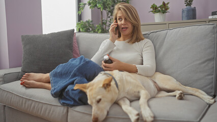 A young caucasian woman converses on a phone, holding medicine, sitting on a couch with her dog at home.