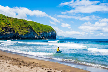 Wall Mural - Unidentified young woman with surfing board walking into ocean water at Saota Barbara beach, Sao Miguel island, Azores, Portugal.