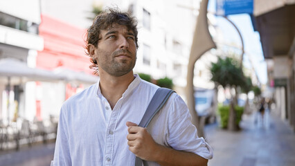 Handsome bearded man with green eyes in casual attire carrying a bag, walking outdoors on a sunny urban street.