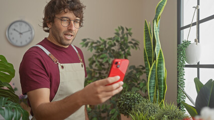 Poster - A young hispanic man with a beard uses a smartphone in an indoor flower shop full of green plants.