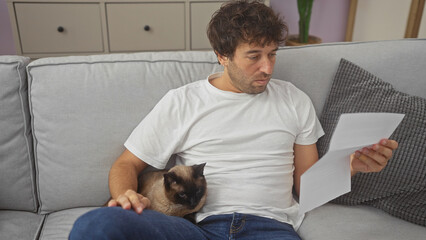 Poster - Young hispanic man reading a document with a siamese cat sitting beside him on a couch indoors.