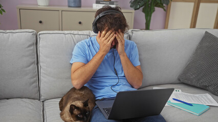 Poster - Stressed man with headphones and laptop sits beside cat on couch in home office interior.