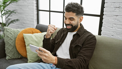 Sticker - A cheerful young man with a beard celebrates a victory at home while holding a smartphone