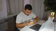 © Krakenimages.com - Young hispanic man in a living room holding a yellow mug and looking at his phone while seated at a table with a laptop
