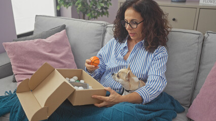 Sticker - A middle-aged hispanic woman sitting on a couch indoors, holding a small basketball, with a curious chihuahua pet beside her, looking at a package.