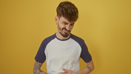 Poster - A young hispanic man with a beard winces in discomfort against a yellow background, suggesting pain or unease in a casual setting.