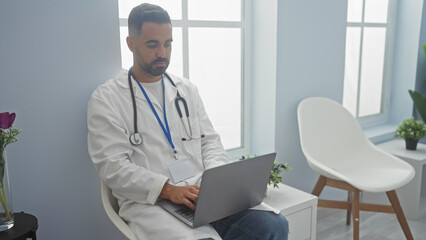 Wall Mural - Hispanic male doctor in white lab coat using laptop in a bright clinic office, embodying professionalism and healthcare.