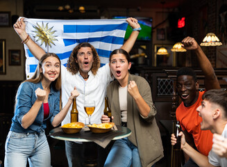 Wall Mural - Multiracial Uruguay sports fans, men and women, supporting their favourite team in bar, raising state flag and screaming chants together.