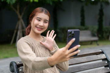 A positive Asian woman is taking a selfie or talking on a video call on a bench in a park.