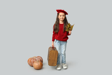 Cute little girl in stylish clothes with autumn leaves, pumpkins and suitcase on grey background