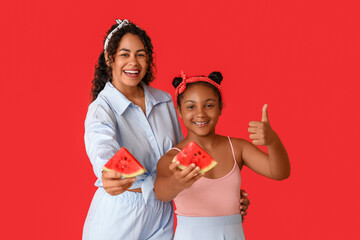 Wall Mural - Beautiful young African-American woman and her cute daughter with pieces of fresh watermelon showing thumb-up gesture on red background