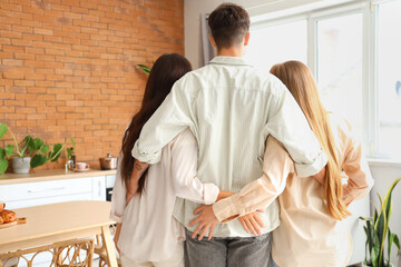 Poster - Young man with two women hugging in kitchen, back view. Polyamory concept