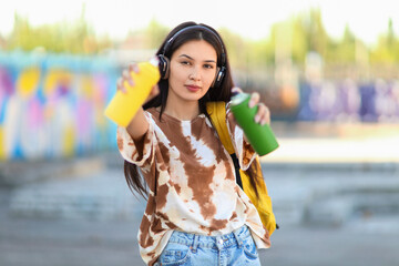 Wall Mural - Young Asian woman in headphones with spray paint cans on street