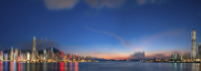 Poster - A panoramic view of Hong Kong, China at dusk with a blurred cityscape and a calm harbor in the foreground.