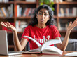© PRASANNAPIX - indian woman in a red t-shirt sitting at a desk with an open book, she looks confused and sad while reading it