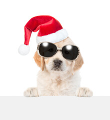 Aufkleber - Cute Golden retriever puppy wearing sunglasses and red santa hat looks above empty white banner. isolated on white background