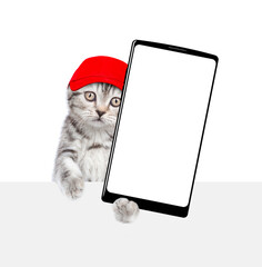 Aufkleber - Cute kitten wearing red cap holds big smartphone with white blank screen in it paw above empty white banner. isolated on white background