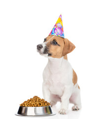 Wall Mural - Jack russell terrier puppy wearing party cap sits with bowl of dry dog food and looks away and up on empty space. isolated on white background