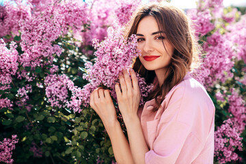 Young beautiful smiling woman in trendy summer clothes. Sexy carefree model posing in the street. Positive model near blooming flower lilac bush beaming. Summertime, pink colours. Red lips