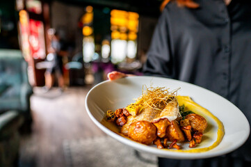 Wall Mural - Close-up of a gourmet dish held by a person, with a blurred restaurant background. The dish features vibrant colors and intricate presentation, highlighting fine dining and culinary artistry.