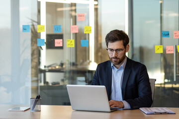 Wall Mural - Focused businessman working on laptop in modern office. Glass wall with colorful sticky notes in background, reflecting collaborative and organized environment. Professional attire suggests corporate