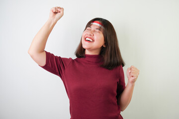 Excited young asian woman wearing Indonesian flag headband, smiling expression, hands raised with clenched fist gesture, isolated over white background. Concept for Indonesian Independence Day.