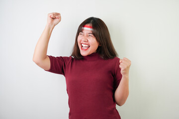 Excited young asian woman wearing Indonesian flag headband, smiling expression, hands raised with clenched fist gesture, isolated over white background. Concept for Indonesian Independence Day.