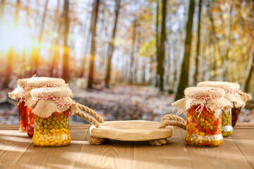 Wall Mural - Autumn pickles of vegetables in glass jars on autumn table with an empty space for products. In the background, an autumn forest trees with yellow and orange leaves.