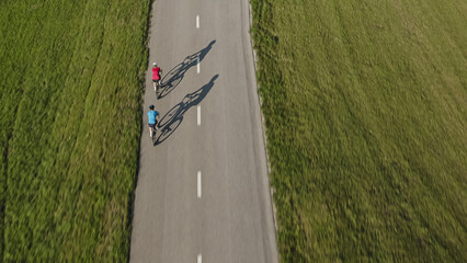 Wall Mural - Two people cycling road bikes, one after the other, on an empty flat roadway between green meadow fields, aerial drone shot.