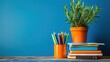 © Ingenious Buddy  - stack of books and colored pencils on a table with an orange plant pot set against a blue wall background, books and pencils, study materials, educational decor