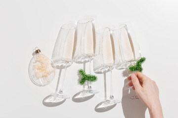 Wall Mural - Minimal New Year flat lay with white wine in champagne glasses,woman hand take one of them, Christmas green tinsel and glass ball as decor. Holiday aesthetic lifestyle, top view, sunlight, shadow