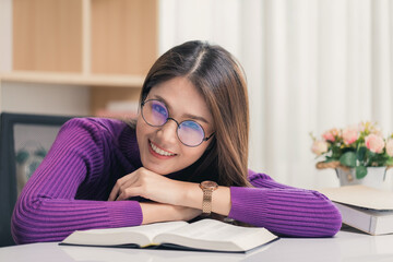 Beautiful young Asian woman wearing glasses is happy while reading a book at home and looking at camera.