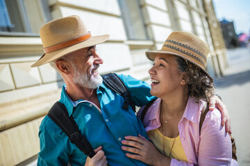 Wall Mural - Senior married couple exploring the city. Hugging each other. They are wearing hats and backpacks.