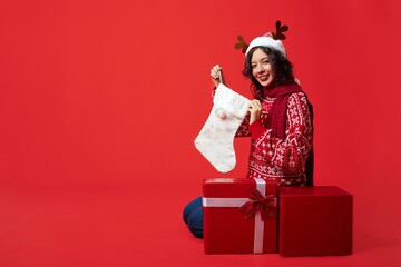 A woman with dark hair and reindeer antlers headband sits on a red gift box next to two more boxes. She wears a striped sweater and jeans, holds a white sock, and smiles.