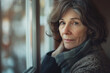 © Bonsales - Middle-aged woman with wavy hair and scarf looks thoughtfully out window. Scene captures contemplation, warmth, and soft indoor light