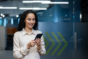 Wall Mural - Businesswoman standing in modern office using smartphone. Woman smiling and engaging with mobile device in professional workspace, illustrating technology, communication, and professional concepts.