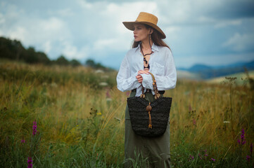 Wall Mural - Fashionable confident woman wearing trendy summer hat, white linen shirt, green maxi skirt, holding black wicker straw bag, posing outdoor. Fashion portrait. Copy, empty space for text
