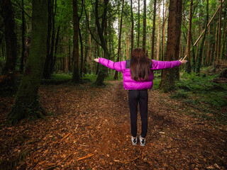 Wall Mural - Young teenager girl in a dark forest park with hands up in the air. Travel and tourism concept. Model is with long dark hair and slim body type wearing colorful jacket and black trousers.