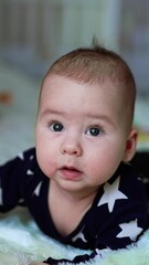 Poster - Beautiful little kid straightened his arms to look up with curiosity. Caucasian child in dark pajamas lies on belly on the bed. Vertical video