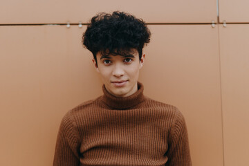 Wall Mural - Fashionable young man with curly hair wearing a brown turtleneck sweater posing against an orange wall with his hands in pockets