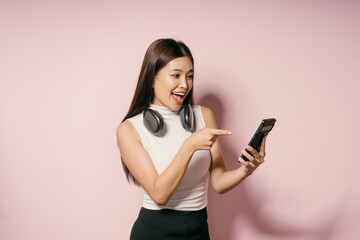 Wall Mural - Young Woman with Headphones Pointing at Smartphone Against Pink Background