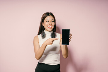 Wall Mural - Young Woman Smiling and Pointing at Smartphone Screen with Headphones Around Neck on Pink Background