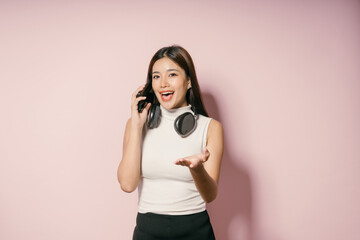 Wall Mural - Young Woman Talking on Phone with Headphones Around Neck Against Pink Background