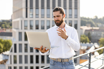 Businessman holding laptop while working outdoors. Person dressed in white shirt stands on building terrace with cityscape background. Concept of remote work business communication modern technology.