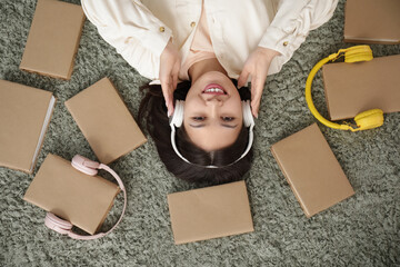 Canvas Print - Beautiful young Asian woman in headphones listening to audiobook with books and lying on carpet at home