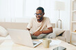© SHOTPRIME STUDIO - African American man working on a laptop in a cozy modern living room, showcasing the lifestyle of a millennial freelancer With a bright smile, he sits comfortably on a sofa, typing away and immersed