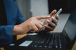 © KANGWANS - A man is using a cell phone while sitting at a desk with a laptop. He is pointing at the screen of the phone