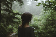 © ALL YOU NEED studio - Woman standing in the forest, back view, on a cloudy day with a misty atmosphere, green trees, wearing a dark blue shirt with a messy hair , blurry background.