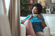 © Dragana Gordic - Happy young African woman sitting on comfortable sofa in living room at home. Smiling pretty girl drinking coffee relaxing on couch looking away through window thinking in modern cozy house.